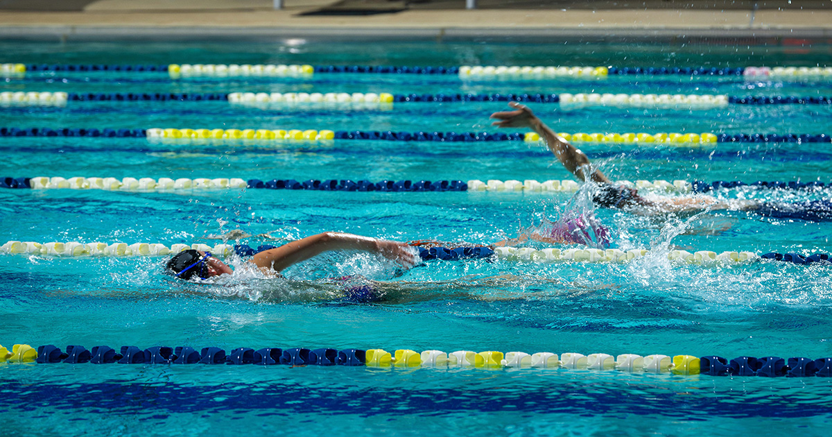 Two people swimming in a pool, captured mid-stroke, with one arm out of the water.