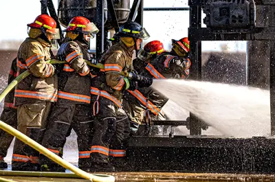 group of firemen in full gear holding a firehouse spraying a fire.