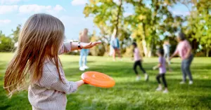 young girl in the foreground throwing a frisbee to family members in an outdoor park while others run and play
