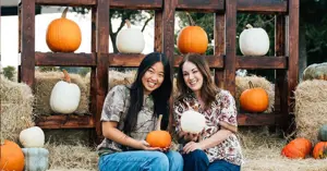 two girls on hay bales holding pumpkins in front of a pumpkin backdrop.