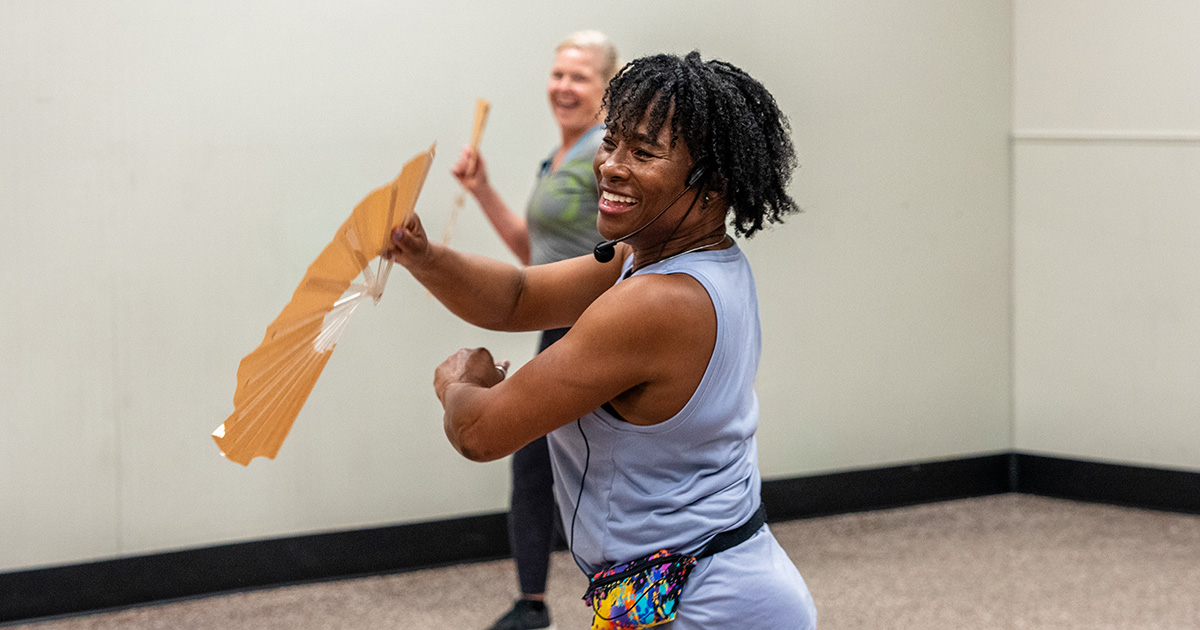 Dance instructor smiling and holding a fan in front of her face with an older woman in the background.