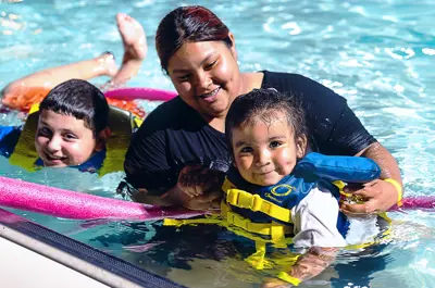 mother holding her toddler who is wearing a colorful lifejacket in the shallows of the city swimming pool
