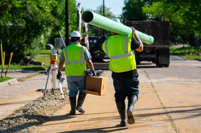 two workers walking toward a streetside worksite carrying a section of water pipe