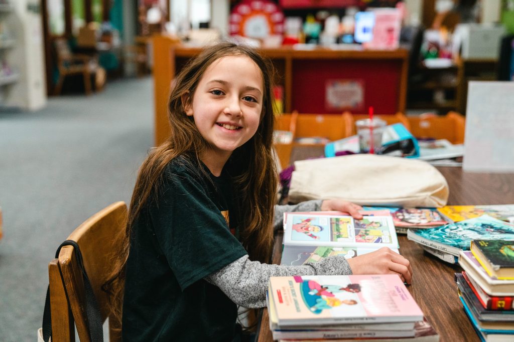 Photo of girl reading at the library smiling at the camera. 