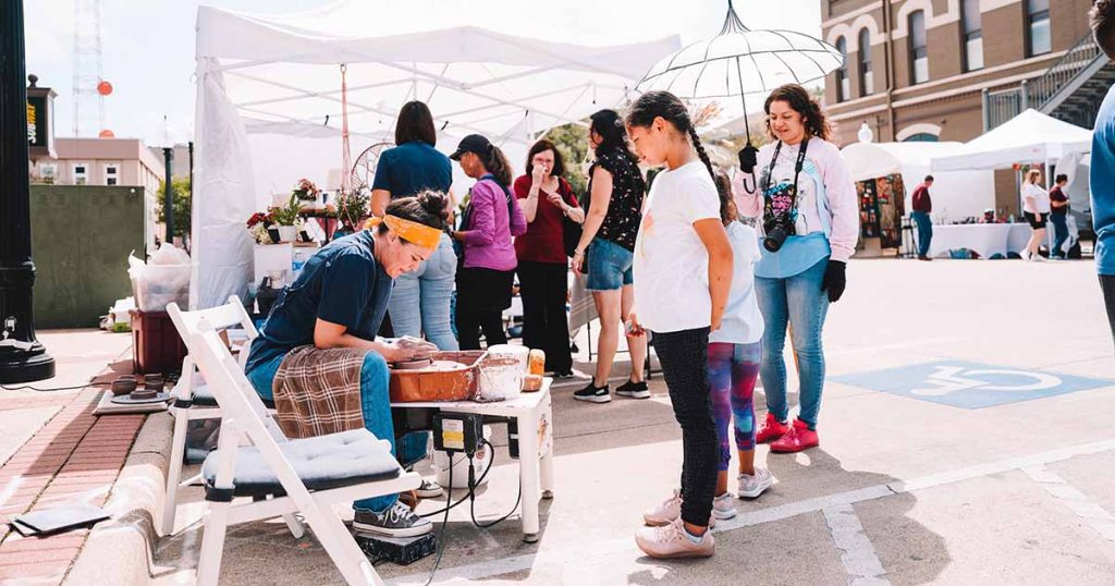 A pottery maker demonstrates spinning a bowl on a pottery wheel to a teenager at the Downtown Bryan Street and Art Fair in 2023.